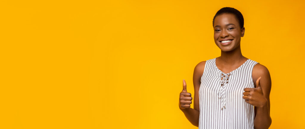 Overjoyed african american girl showing thumbs up to free space, widely smiling, yellow studio background