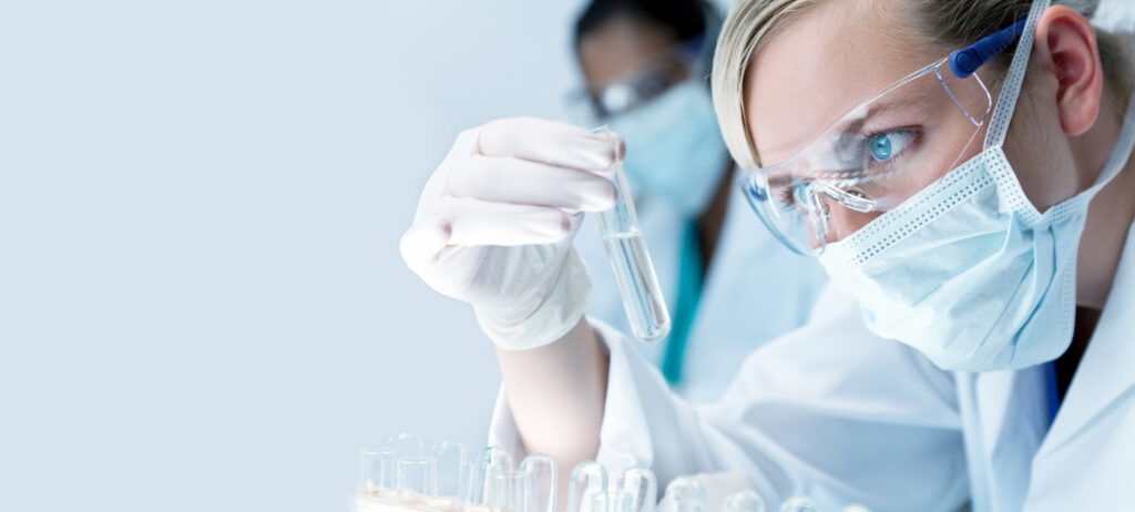 Panorama female woman medical or scientific researcher or doctor wearing a face mask looking at a test tube in a laboratory with her Asian colleague out of focus behind her. Panoramic web banner.