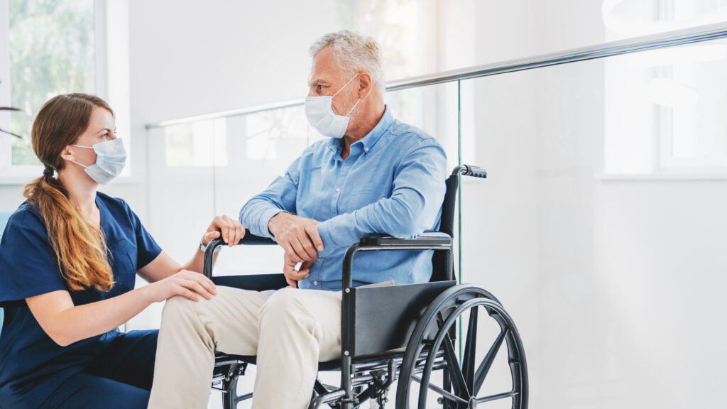 Caucasian nurse taking care of senior male patient on wheelchair in hospital. Young woman and old man wearing face mask for protection of covid 19 pandemic