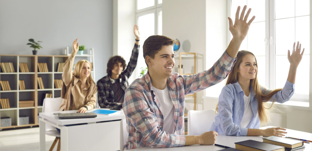 Group of enthusiastic male and female high school or college students sitting at desks and raising hands in class. Pupils voting, willing to answer question or eager to take part in classroom activity