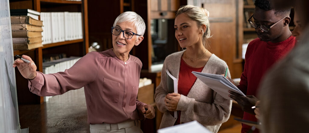 Professor helping student in library at college. Group of multiethnic students taking an active part in a lesson while standing in library. Mature woman teacher and university student discussing over math formulas on whiteboard.