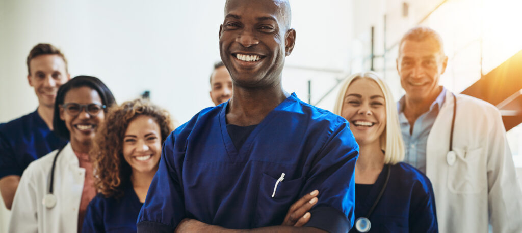 Young African male doctor smiling while standing in a hospital corridor with a diverse group of staff in the background